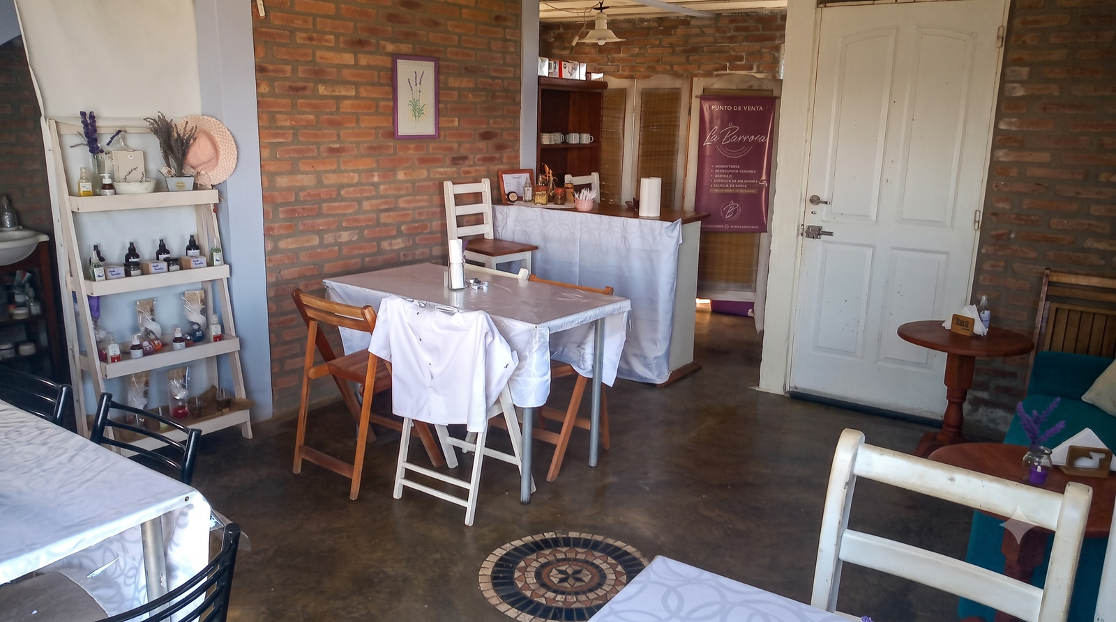 Interior de MAMU Casa de Té — mesas con mantel blanco, pared de ladrillo, estantería con productos y mostrador
